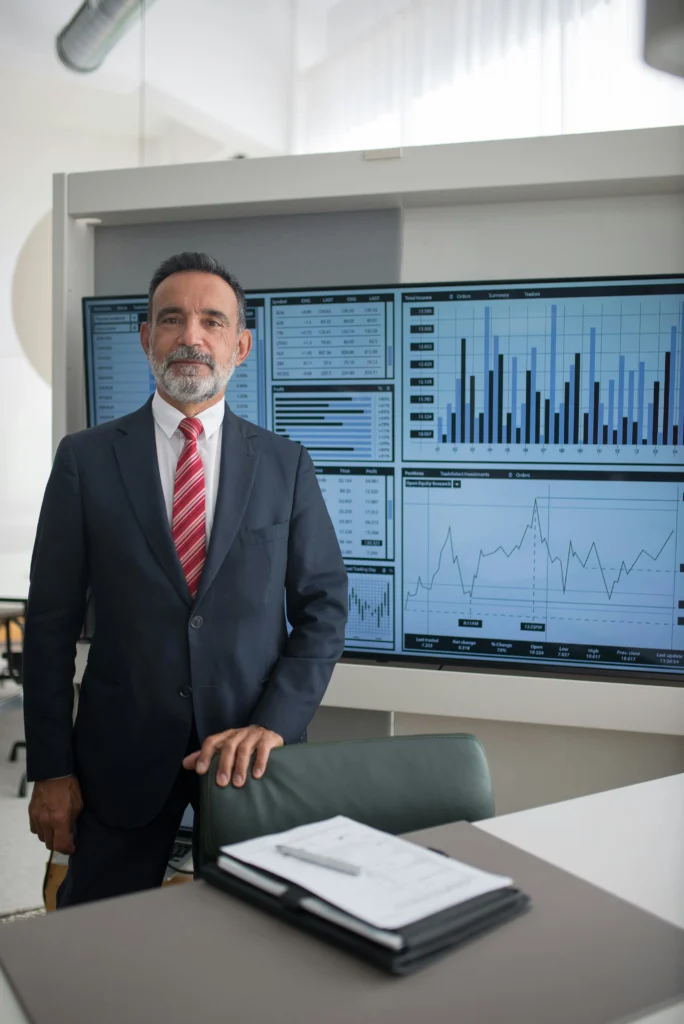 A confident businessman in a formal suit standing in front of a large screen displaying financial charts and market data in a modern office setting.
